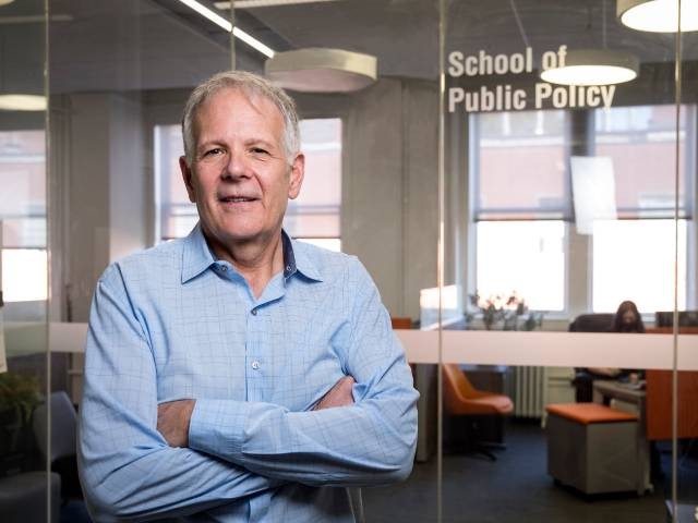 Brent Steel standing in a blue dress shirt in front of a glass wall that reads "School of Public Policy"